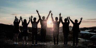 En grupp människor sträcker upp armarna i luften, de står vända mot solnedgång